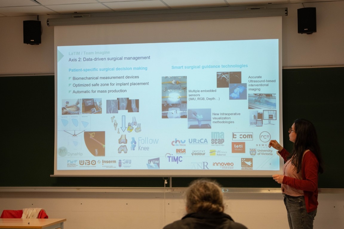 Intervenante présentant une diapositive projetée dans une salle de cours ou de conférence, portant sur le management chirurgical basé sur les données et les technologies de guidage chirurgical intelligent, avec logos de partenaires académiques et institutionnels affichés à l’écran.